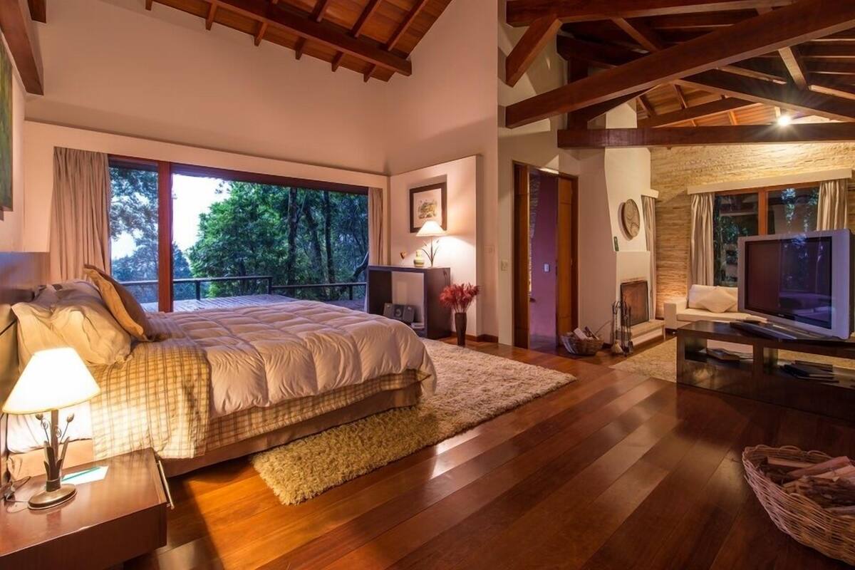 Une chambre spacieuse avec un grand lit, un plafond en bois et une vue sur les arbres par une grande fenêtre.
