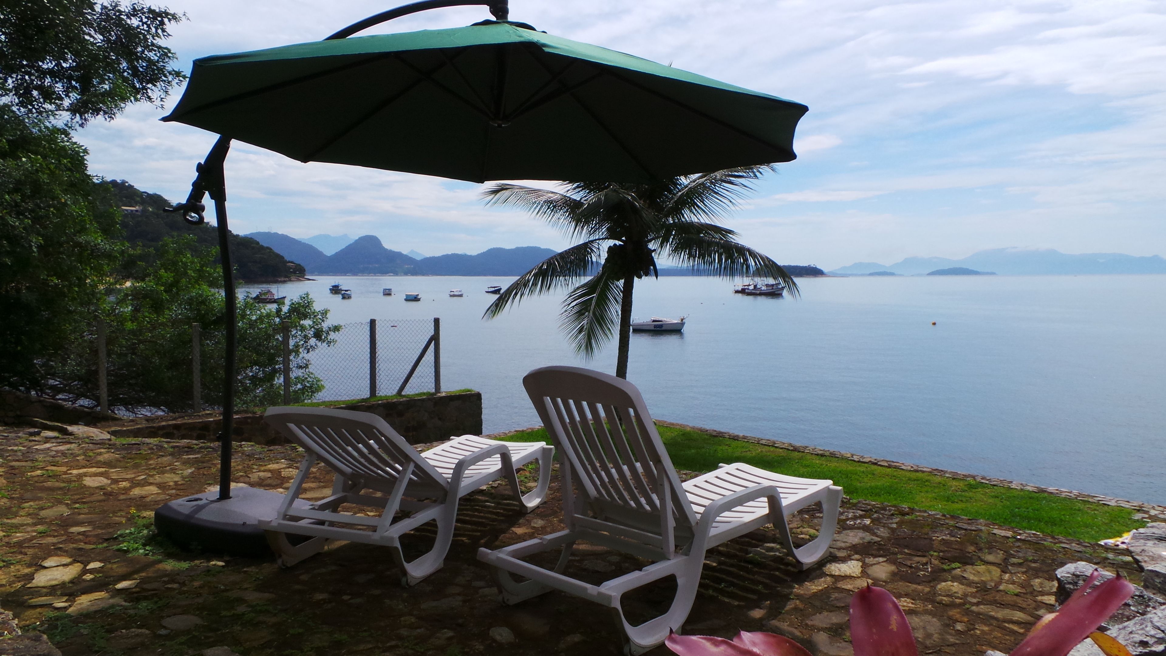A terrace with two white plastic chairs and a green umbrella, overlooking a calm body of water with boats and distant mountains.