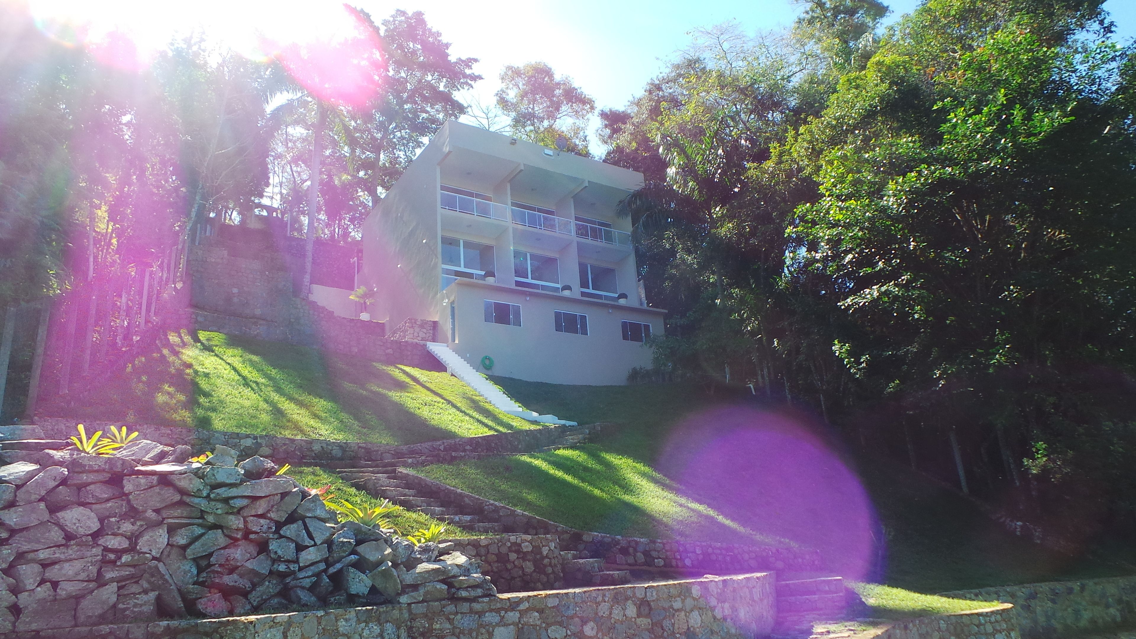 A two-story building with a flat roof and multiple windows, surrounded by lush greenery and a stone wall in the foreground.