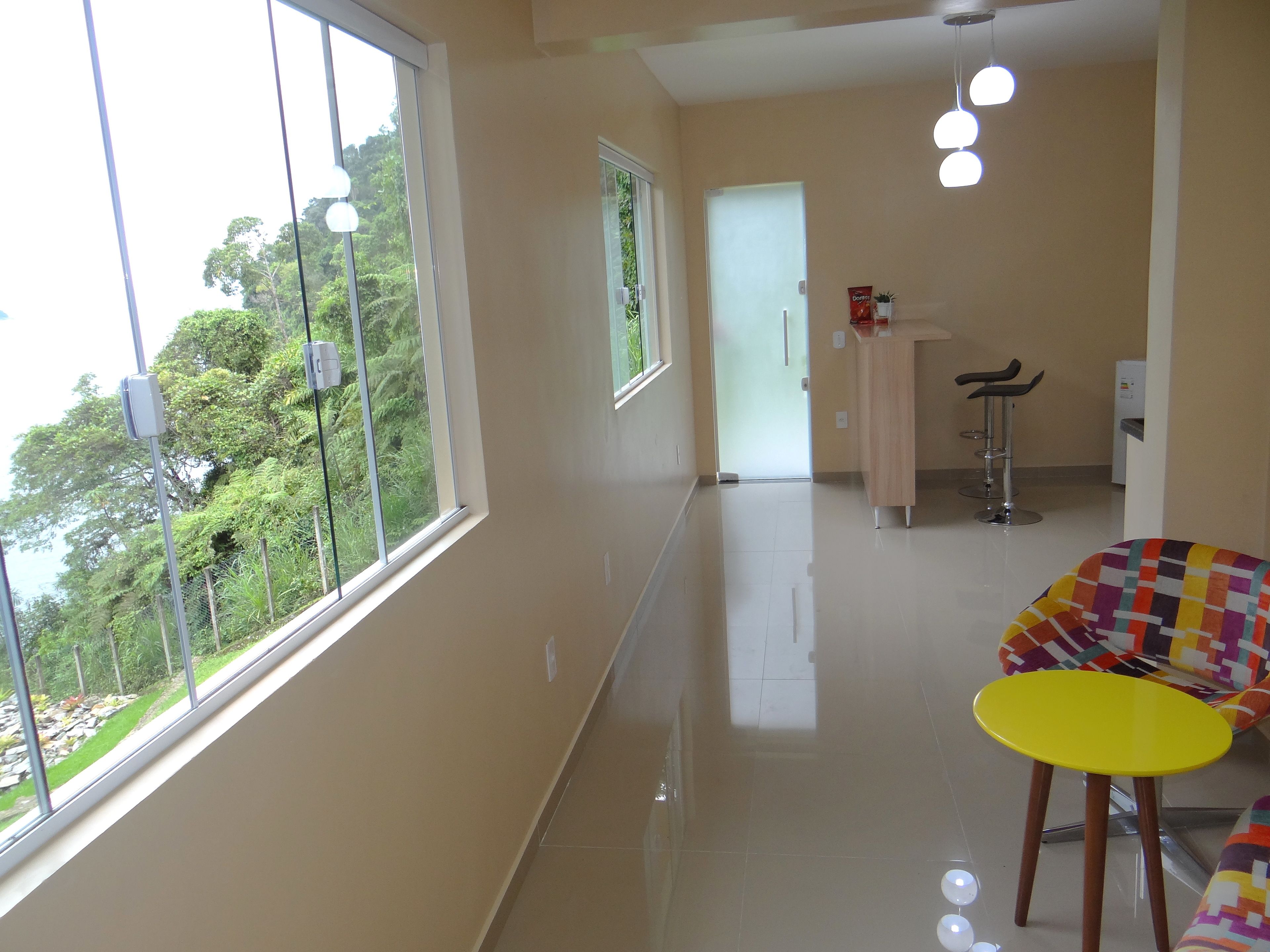Habitación de hotel con ventanas amplias, una mesa amarilla, una silla colorida y una zona de kitchenette.