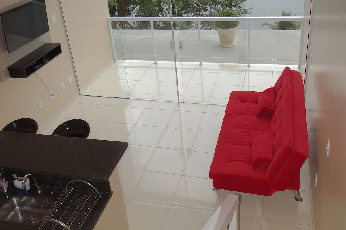 A modern kitchen with a black countertop, a built-in oven, and a sink. A red sofa is placed on a balcony with a view of a body of water and greenery.