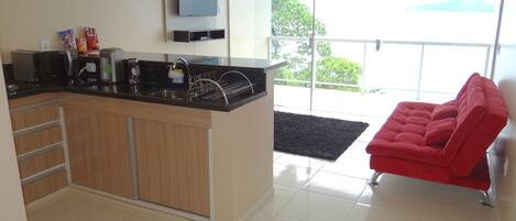 A modern kitchen with a built-in oven, microwave, and coffee maker. There is a red chair and a black rug in the living area.