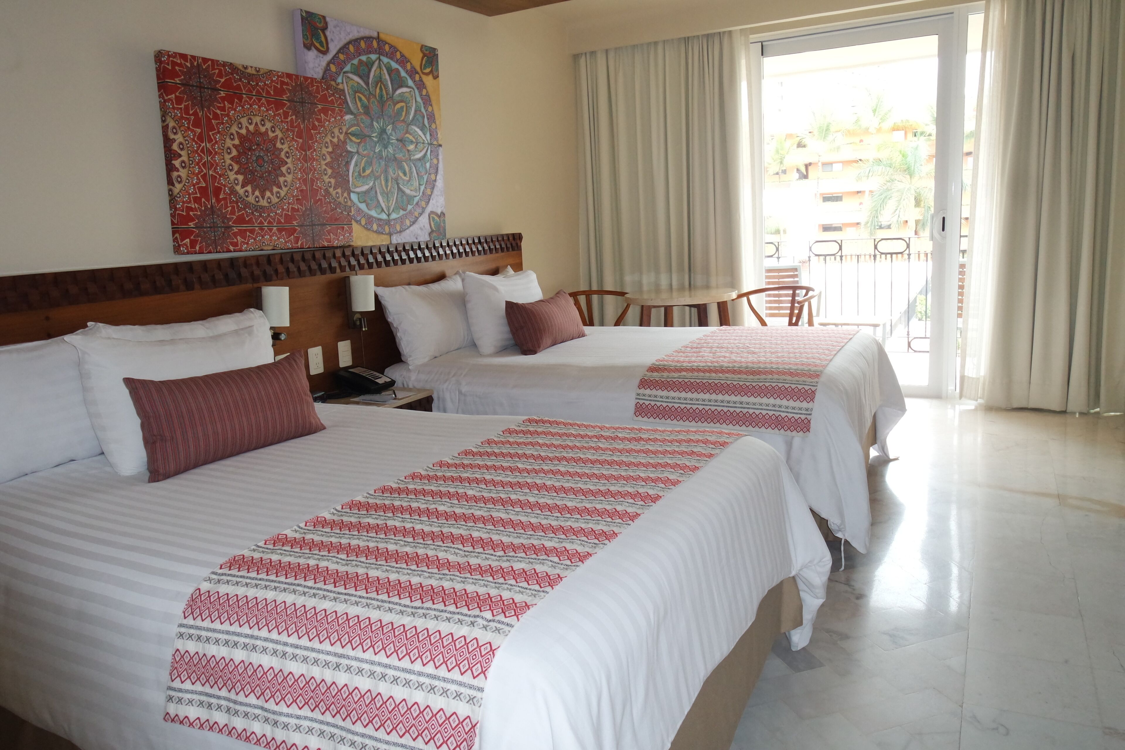 A hotel room with two beds, a wooden headboard, a table with chairs, and a view of the outside balcony.