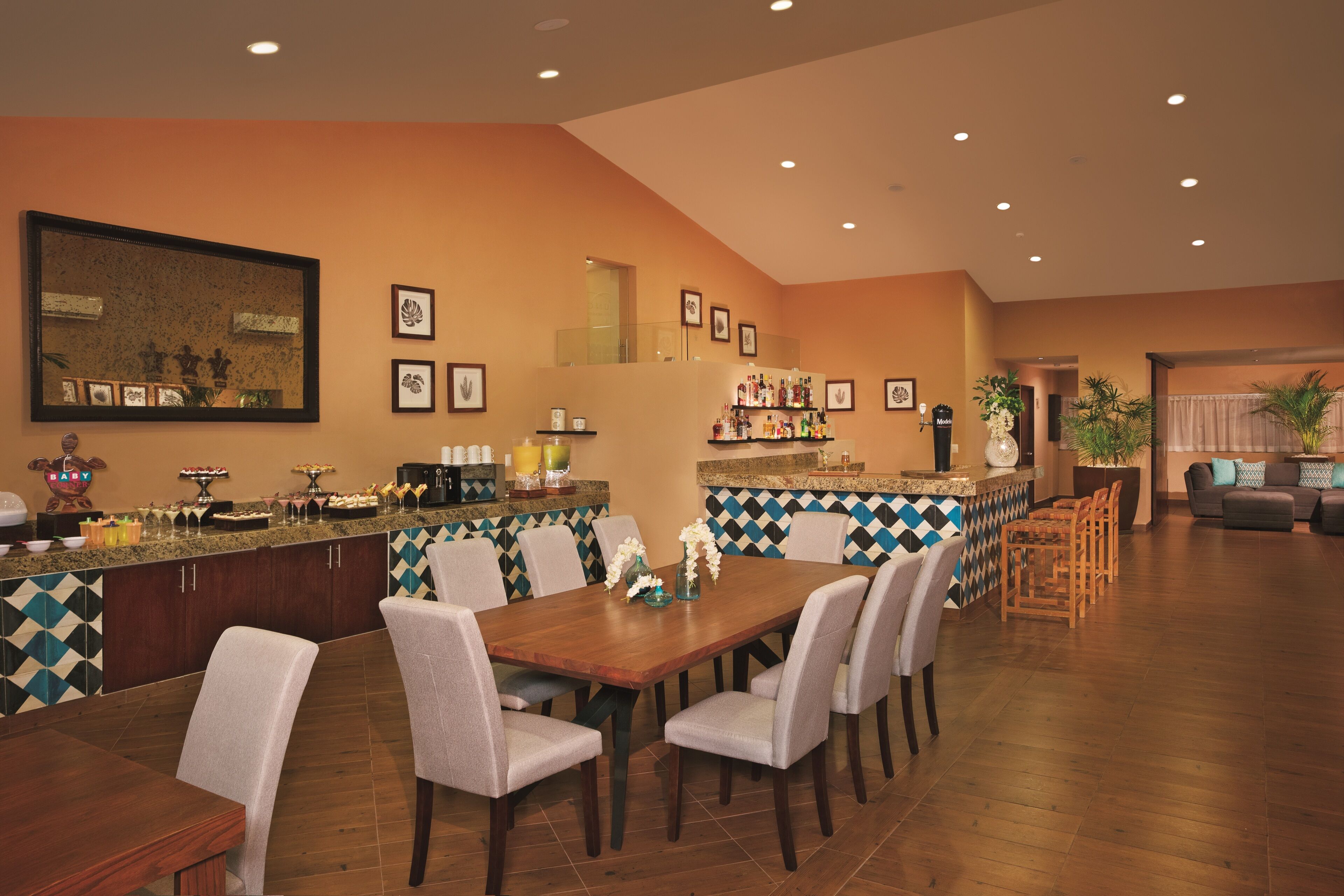 Un comedor amplio con una larga mesa de madera y numerosas sillas, una zona de cocina con barra, y un espacio de estar con sofá y sillones.