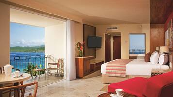 Habitación de hotel con balcón, cama, televisión, zona de comedor y vista al océano.