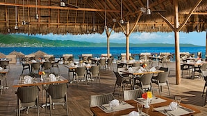 An outdoor dining area with thatched roofs, wooden tables, and chairs, overlooking a coastal view.