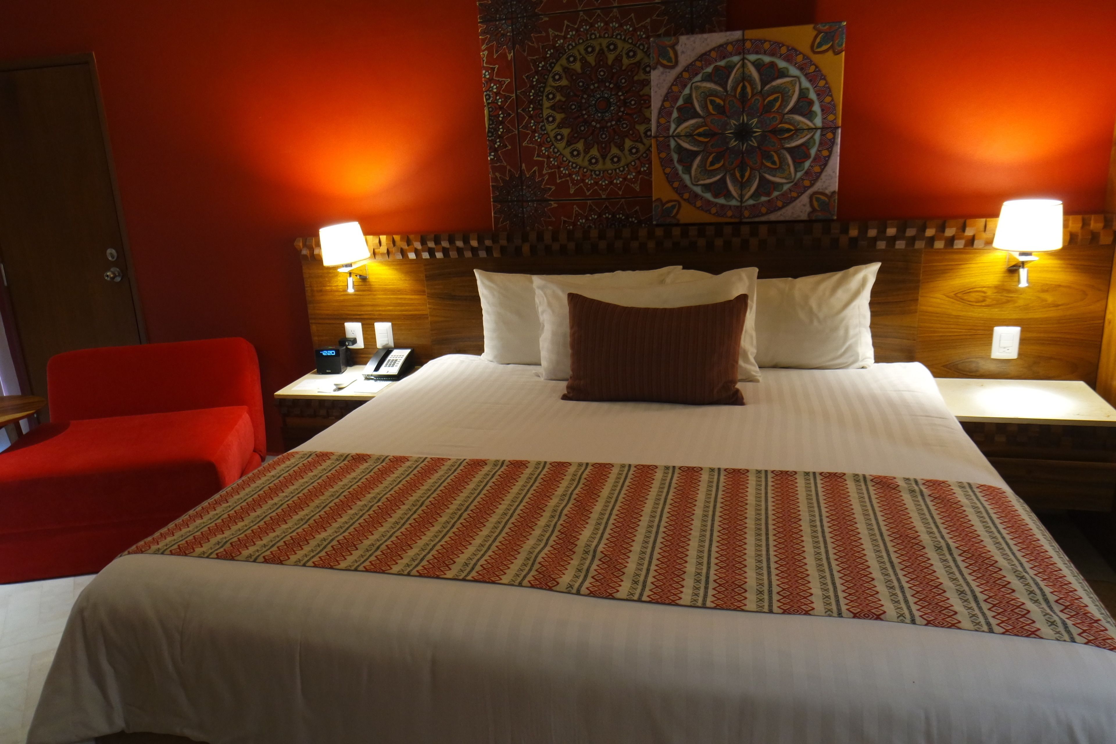 A hotel room with a large bed, two bedside tables, a red chair, and a decorative tapestry on the wall.