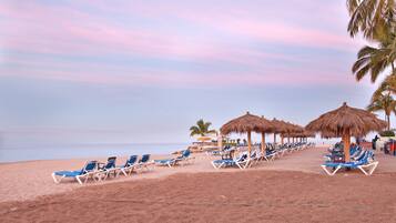 Una playa con hileras de sillas azules, sombrillas de paja, palmeras y un mar tranquilo.