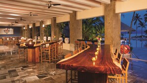 A long wooden dining table with chairs, a bar area, and a view of a pool at dusk.