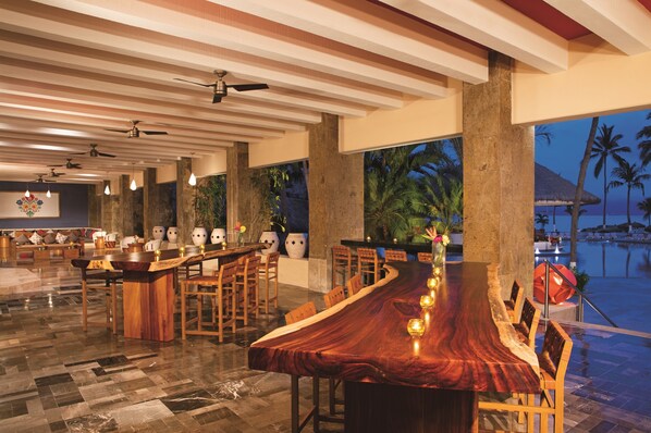 A long wooden dining table with chairs, a bar area, and a view of a pool at dusk. - Sunscape Puerto Vallarta Resort & Spa All Inclusive (Puerto Vallarta)