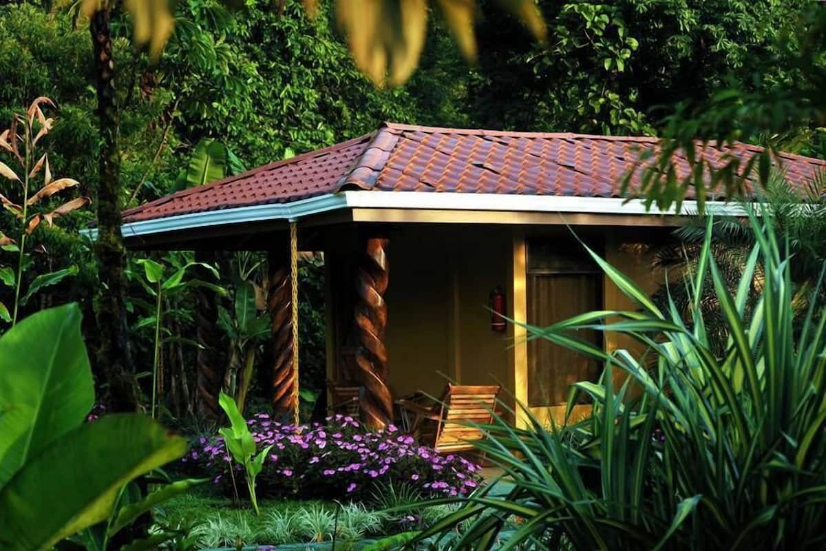 A small, rustic building with a tiled roof, surrounded by lush greenery and flowering plants.