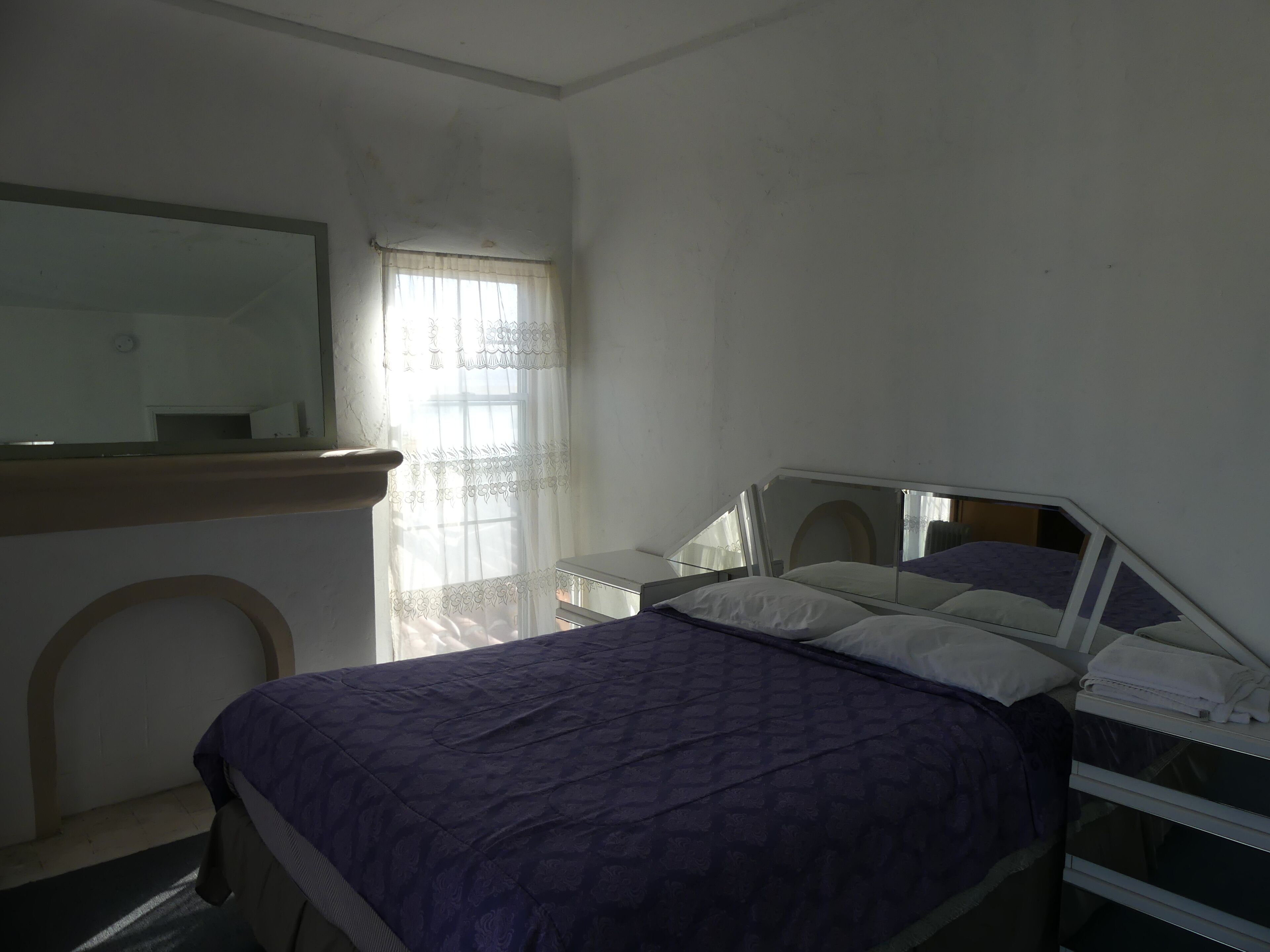 Une chambre à coucher avec un lit, une table de chevet, un miroir et une fenêtre avec un rideau.