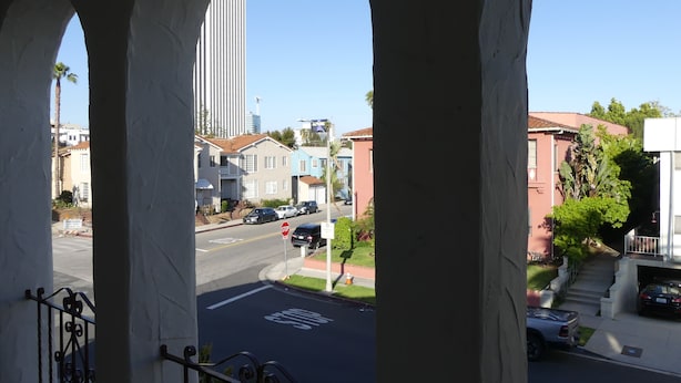 Une vue depuis un balcon, donnant sur une rue avec des immeubles d’habitation, un panneau de signalisation et un ciel bleu dégagé.