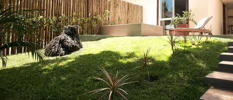 A modern outdoor area with a wooden fence, a grassy lawn, a small tree stump, and a chair near a building entrance.