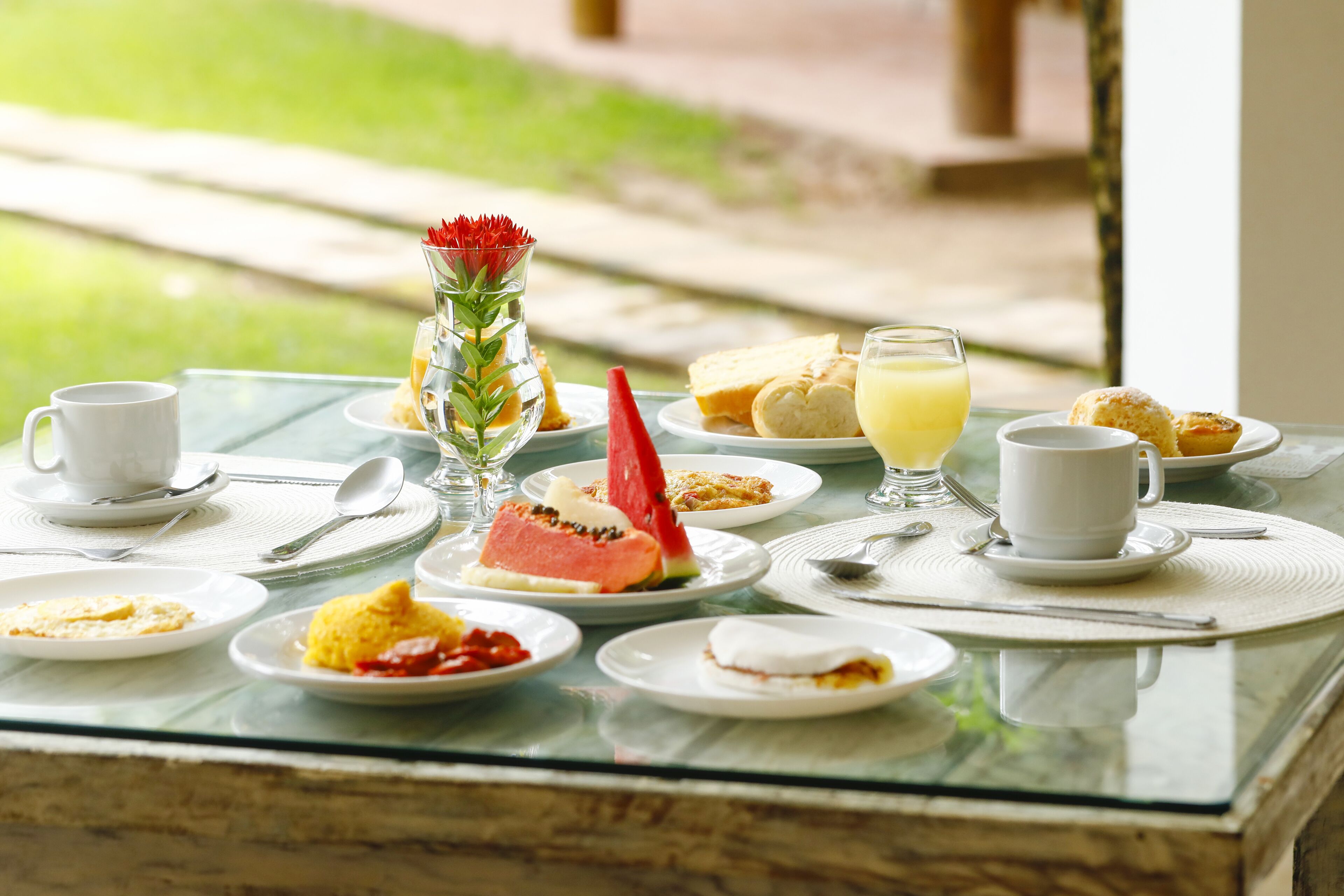 Una mesa de desayuno con diversos platos, un jarrón con una flor roja y un vaso de jugo de naranja.