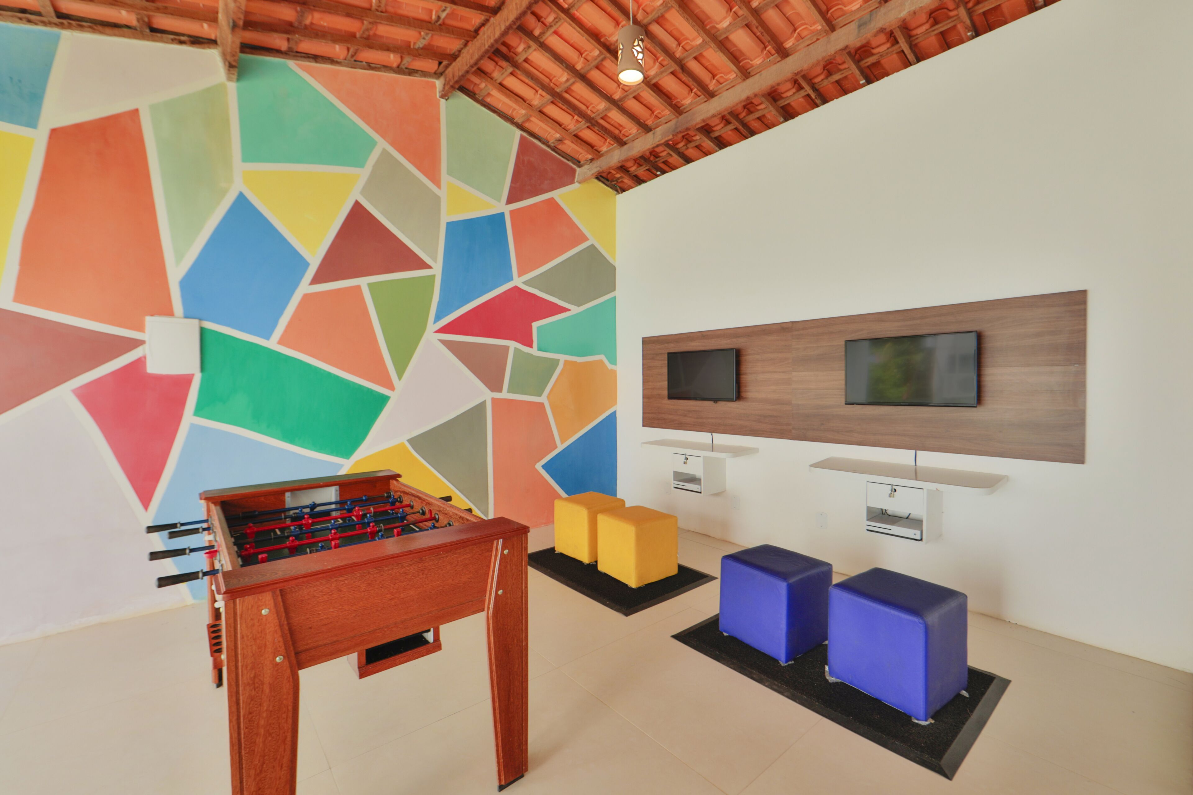 A room with a colorful geometric wall, a wooden foosball table, a TV mounted on the wall, and two blue cushioned seating areas.