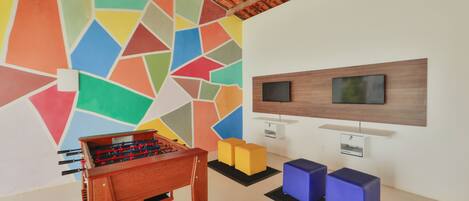A room with a colorful geometric wall, a wooden foosball table, a TV mounted on the wall, and two blue cushioned seating areas.