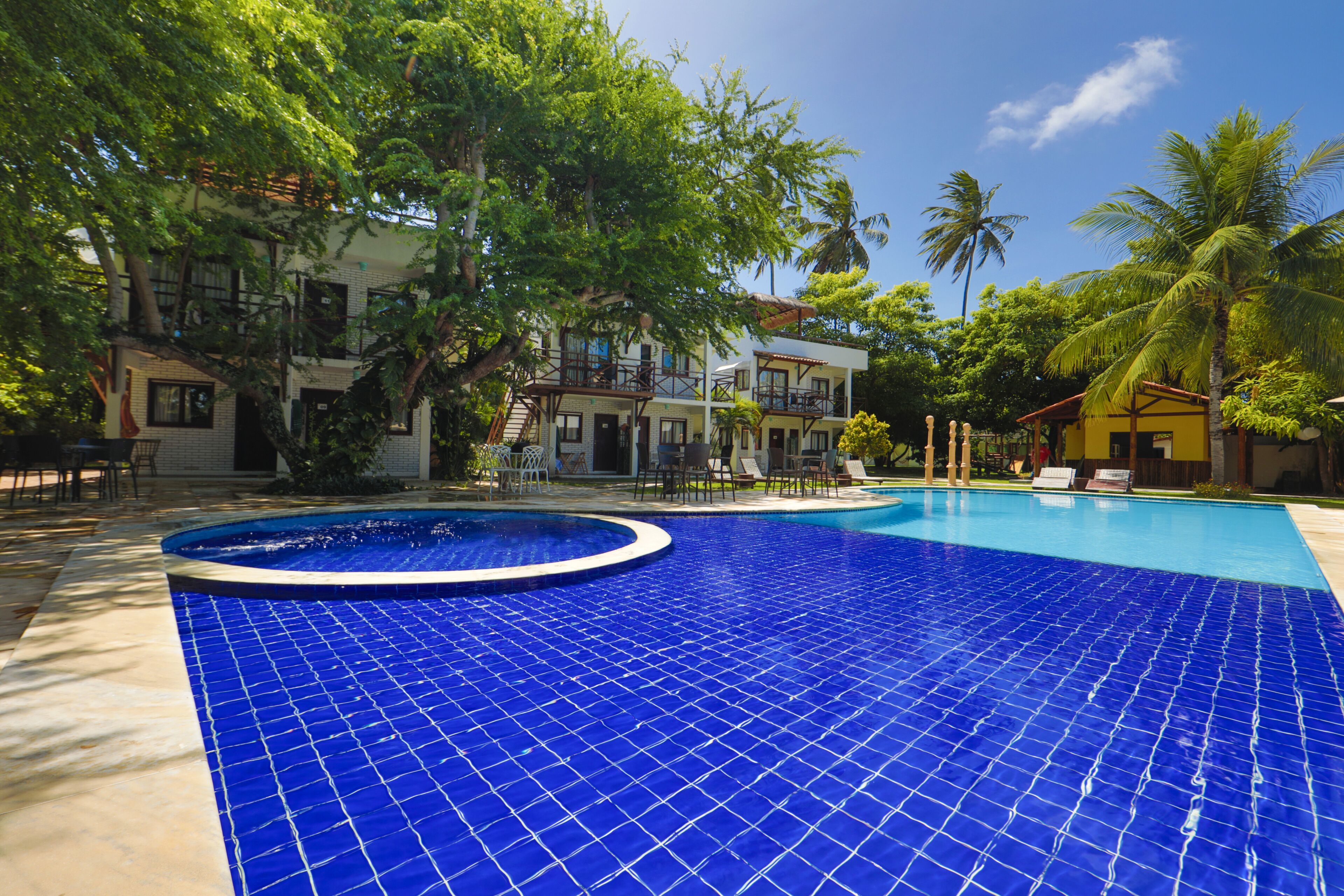 A pool with blue tiles, surrounded by palm trees and a building with multiple balconies.