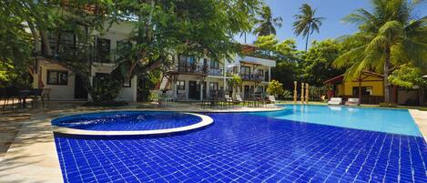 A pool with blue tiles, surrounded by palm trees and a building with multiple balconies.