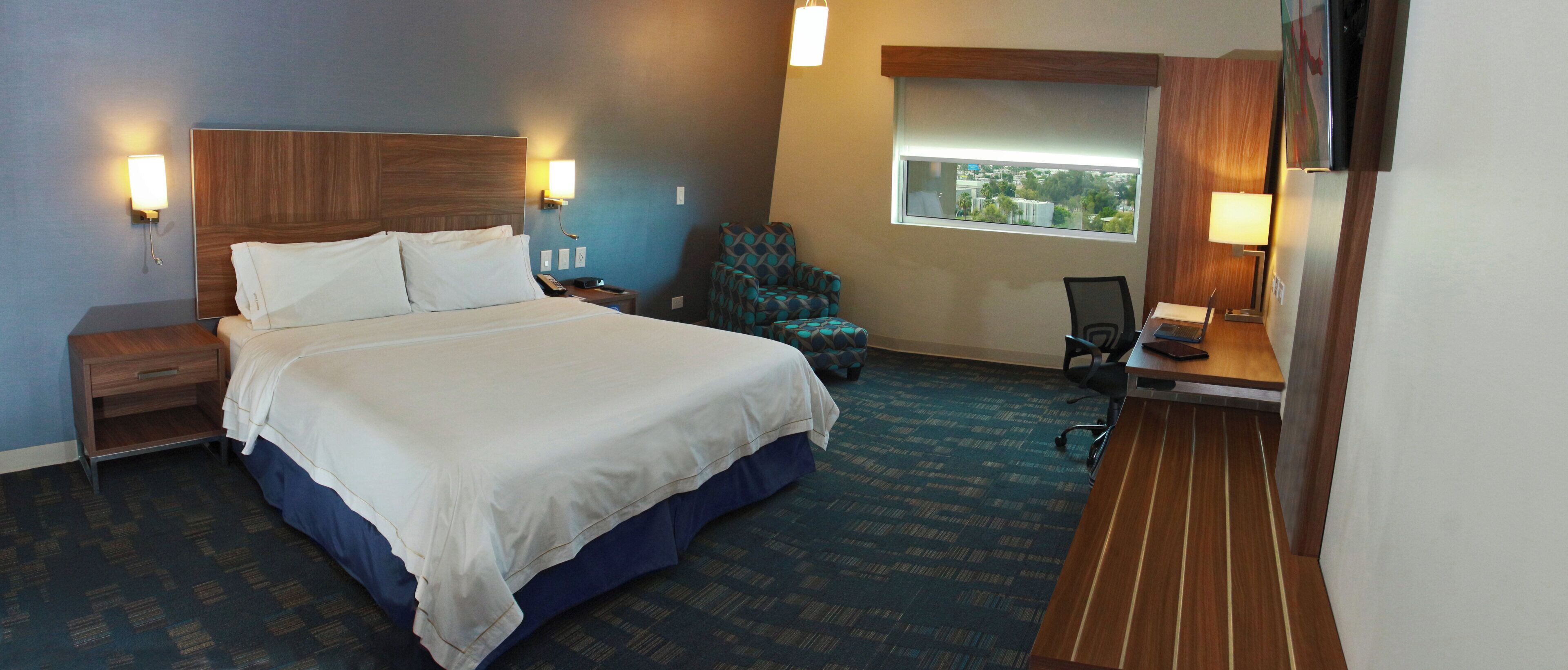 A hotel room with a large bed, a desk with a laptop, a chair, and a window with a view.