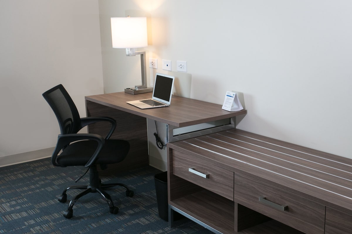 Habitación de hotel con un escritorio de madera, una computadora portátil, una silla, una lámpara y un cesto de basura.