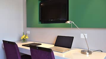 A modern office space with a white desk, a laptop, a tablet, a TV mounted on a green wall, a desk lamp, and purple chairs.
