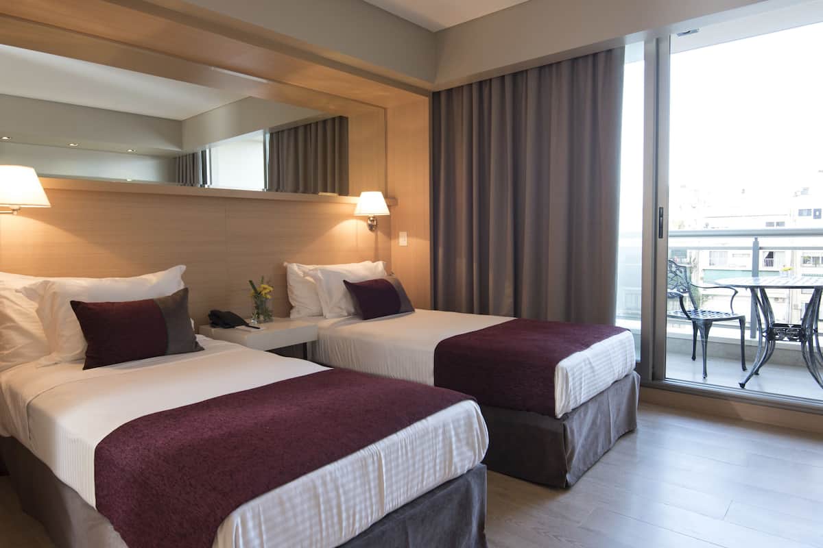 Quarto de hotel com duas camas, uma mesinha, um telefone e varanda com mesa e cadeiras.