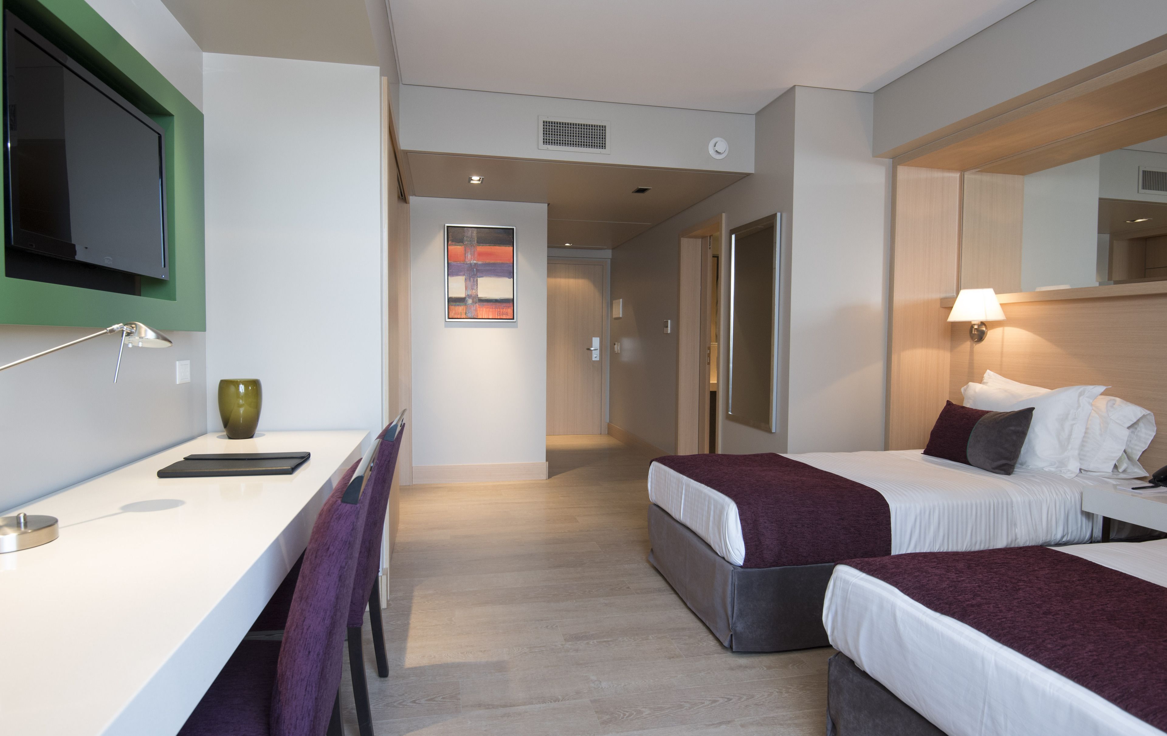 A hotel room with two beds, a desk with a lamp, a TV mounted on the wall, and a painting on the wall.