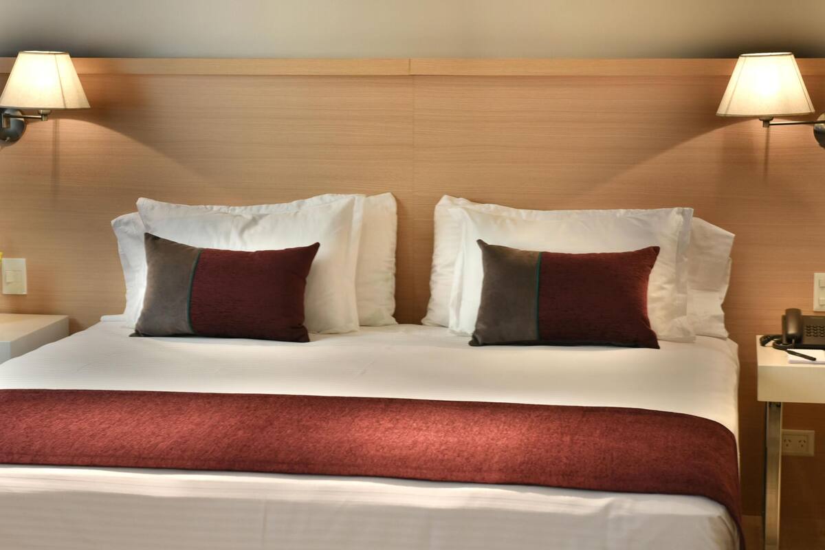 Quarto de hotel com uma cama grande, duas luminárias de cabeceira, mesa de apoio com telefone e um vaso de flores.