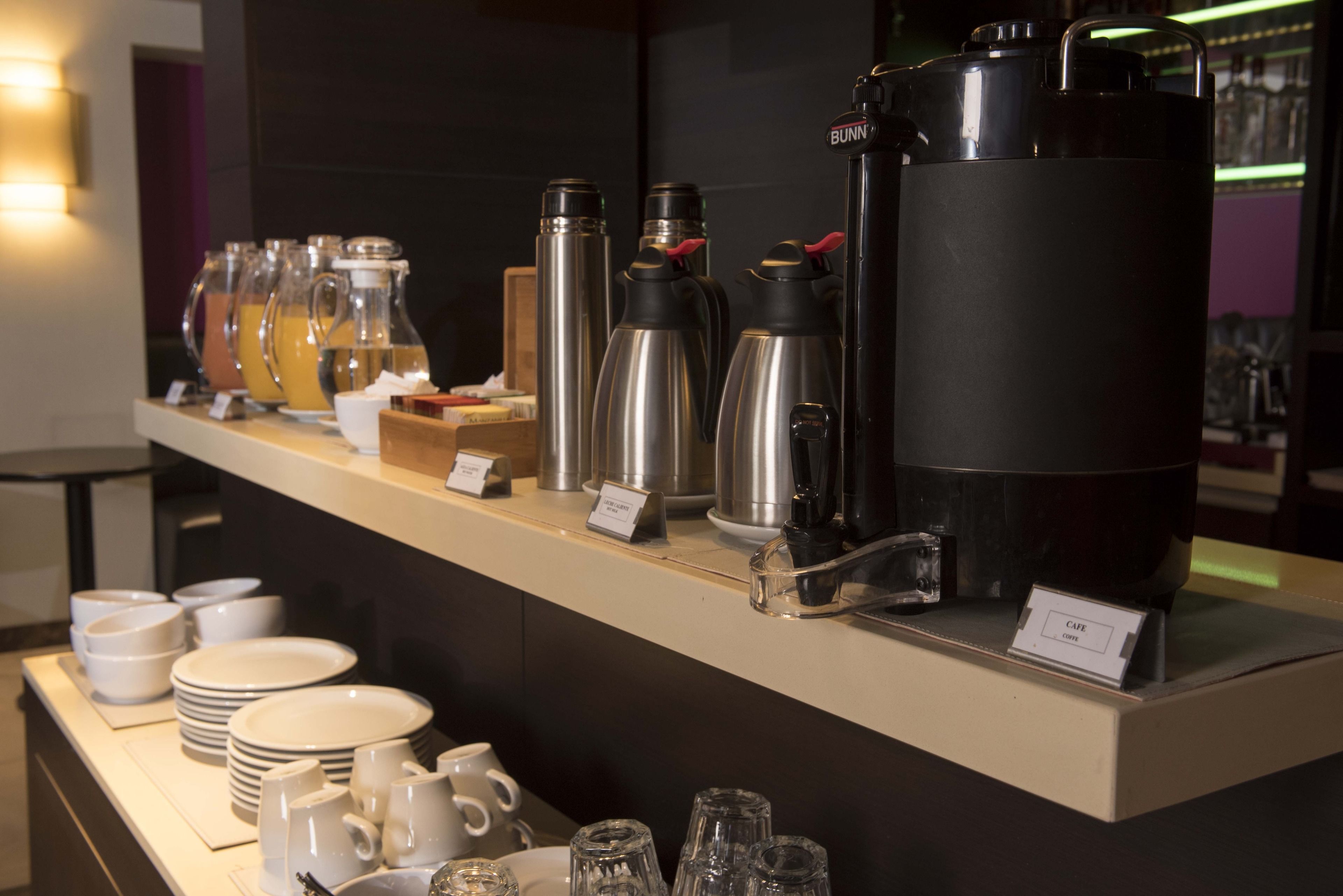 Un comptoir de petit-déjeuner avec différents distributeurs de café et de thé, une machine à café, ainsi que de nombreuses assiettes et tasses blanches.
