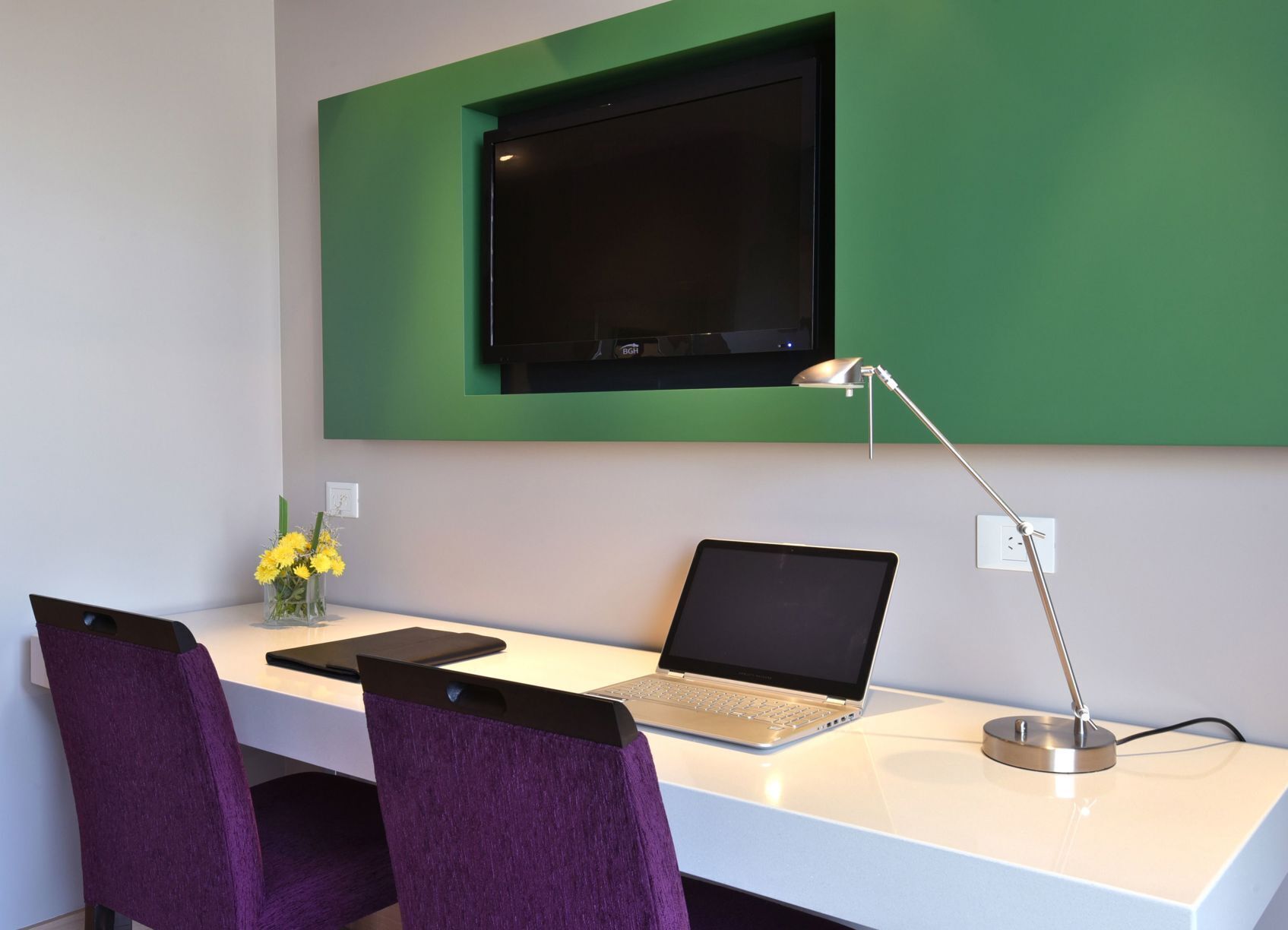 A modern workspace with a laptop, a desk lamp, and a TV mounted on a green wall.