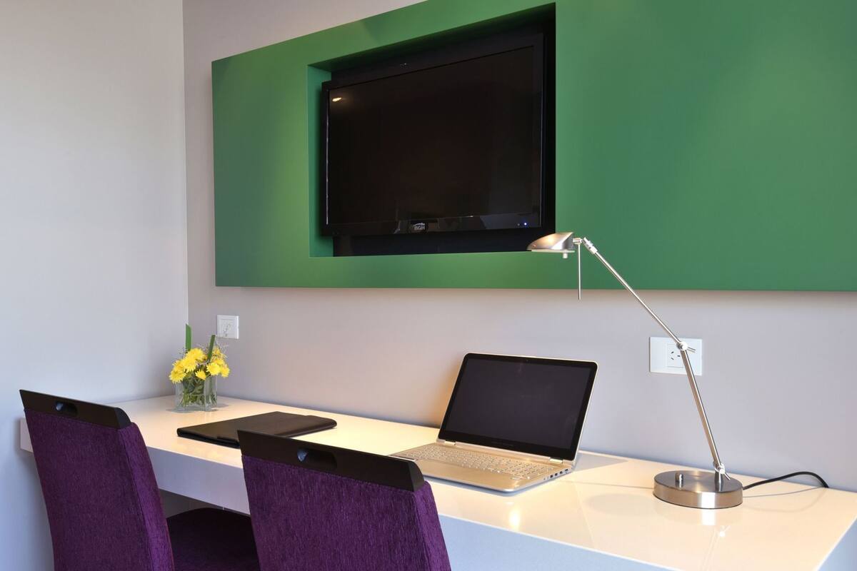 Um espaço de trabalho moderno com um laptop, uma luminária de mesa e uma TV fixada em uma parede verde.