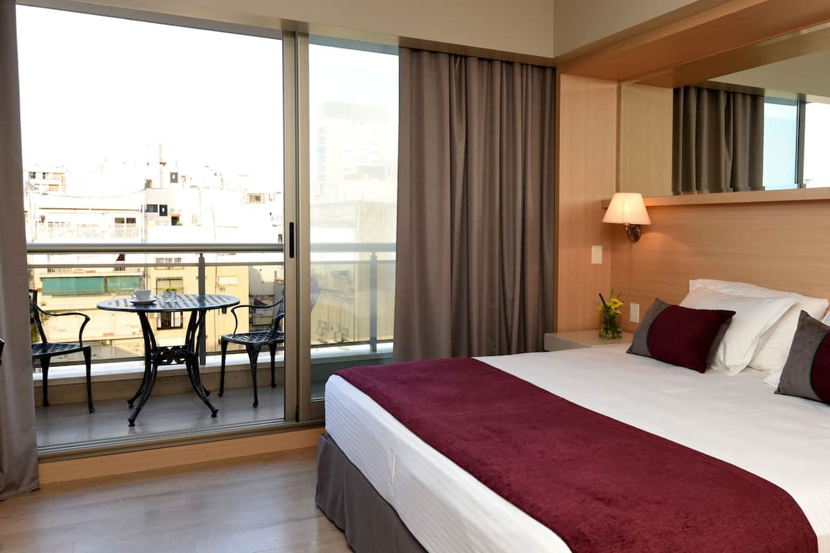 Quarto de hotel com uma cama grande, varanda com mesa e cadeiras, e vista para edifícios.
