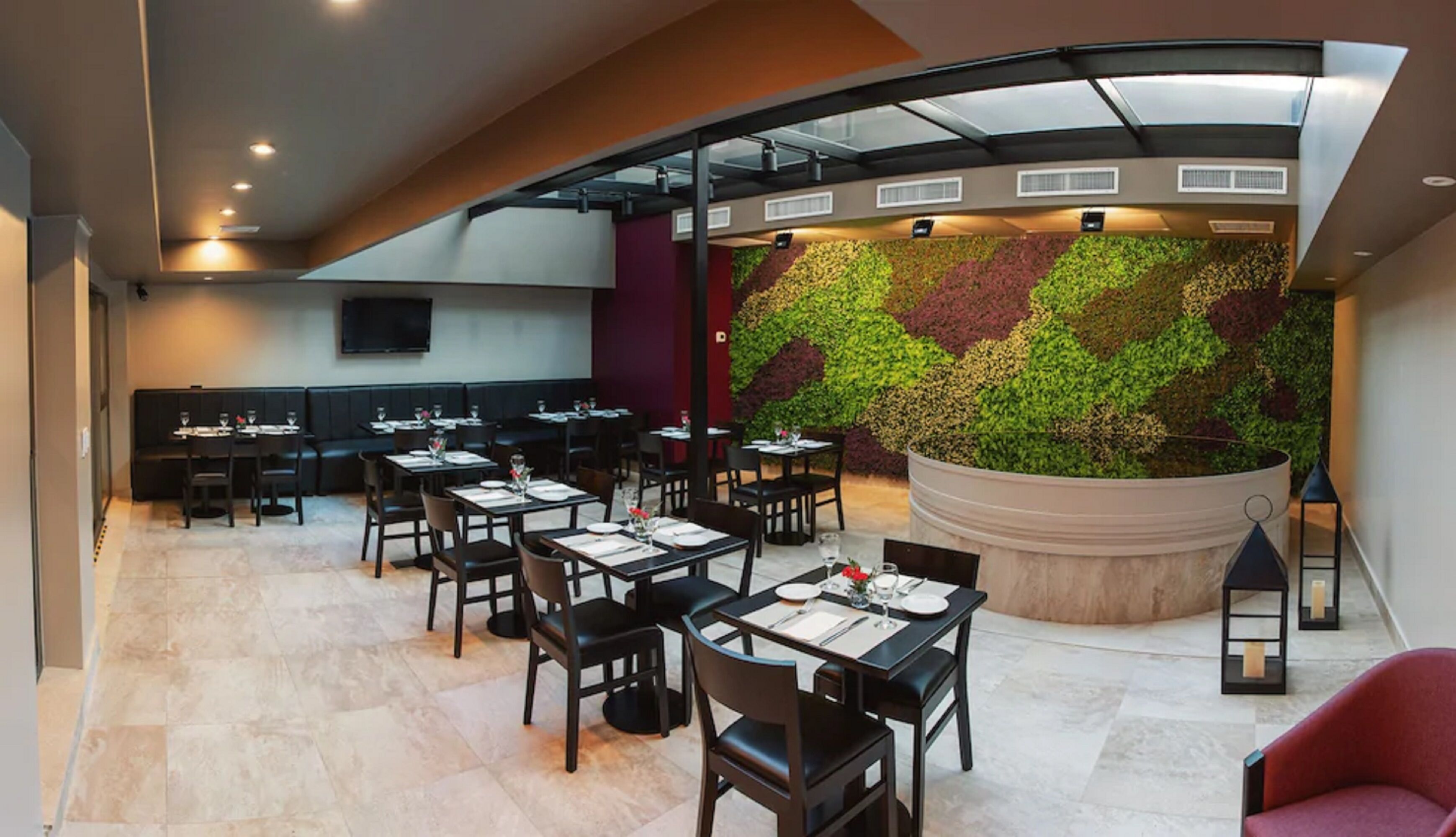 Un restaurante con una decoración moderna, que presenta un techo curvo, una pared verde con un diseño de hojas, y diversas opciones de asientos, incluyendo mesas y cabinas.