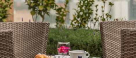 A wicker chair, a glass table with a plate of pastries, a cup, and a glass with a flower.