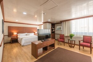 Une chambre d’hôtel avec une tête de lit en bois, un lit, un téléviseur posé sur un meuble en bois, un bureau avec une chaise et une fenêtre avec des rideaux.