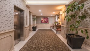A hallway with patterned wallpaper, a brown carpet, an elevator, a potted plant, and a painting on the wall.