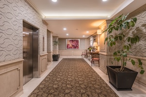 A hallway with patterned wallpaper, a brown carpet, an elevator, a potted plant, and a painting on the wall.