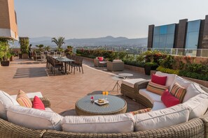 A rooftop patio with wicker furniture, a round table, and a view of the city and mountains.