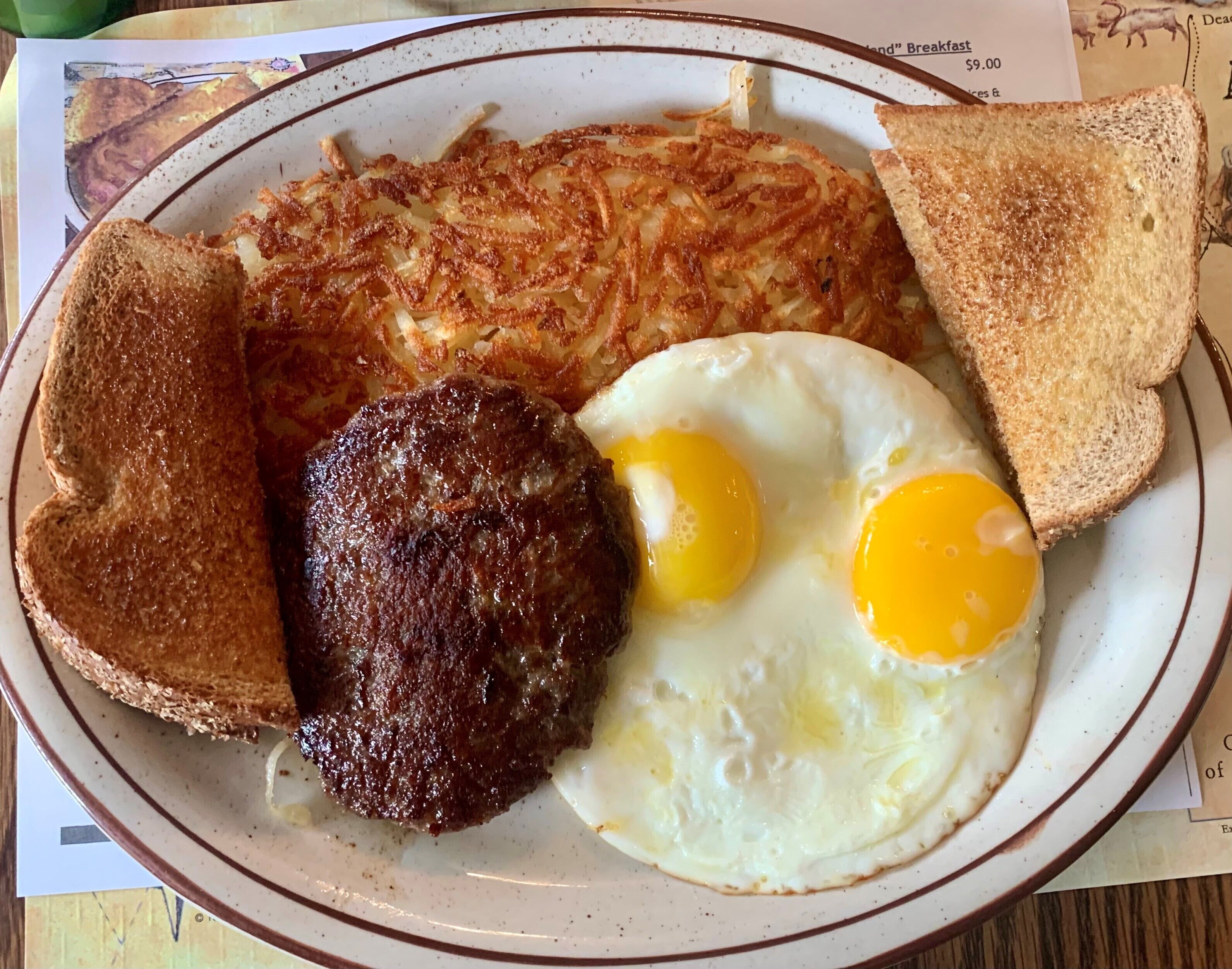 A breakfast plate with hash browns, two fried eggs, a sausage patty, and two slices of toast.
