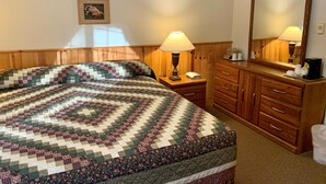 A hotel room with a large bed, a nightstand with a lamp, a dresser with drawers, a mirror, and a framed picture on the wall.