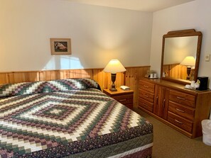 A hotel room with a large bed, a nightstand with a lamp, a dresser with drawers, a mirror, and a framed picture on the wall.