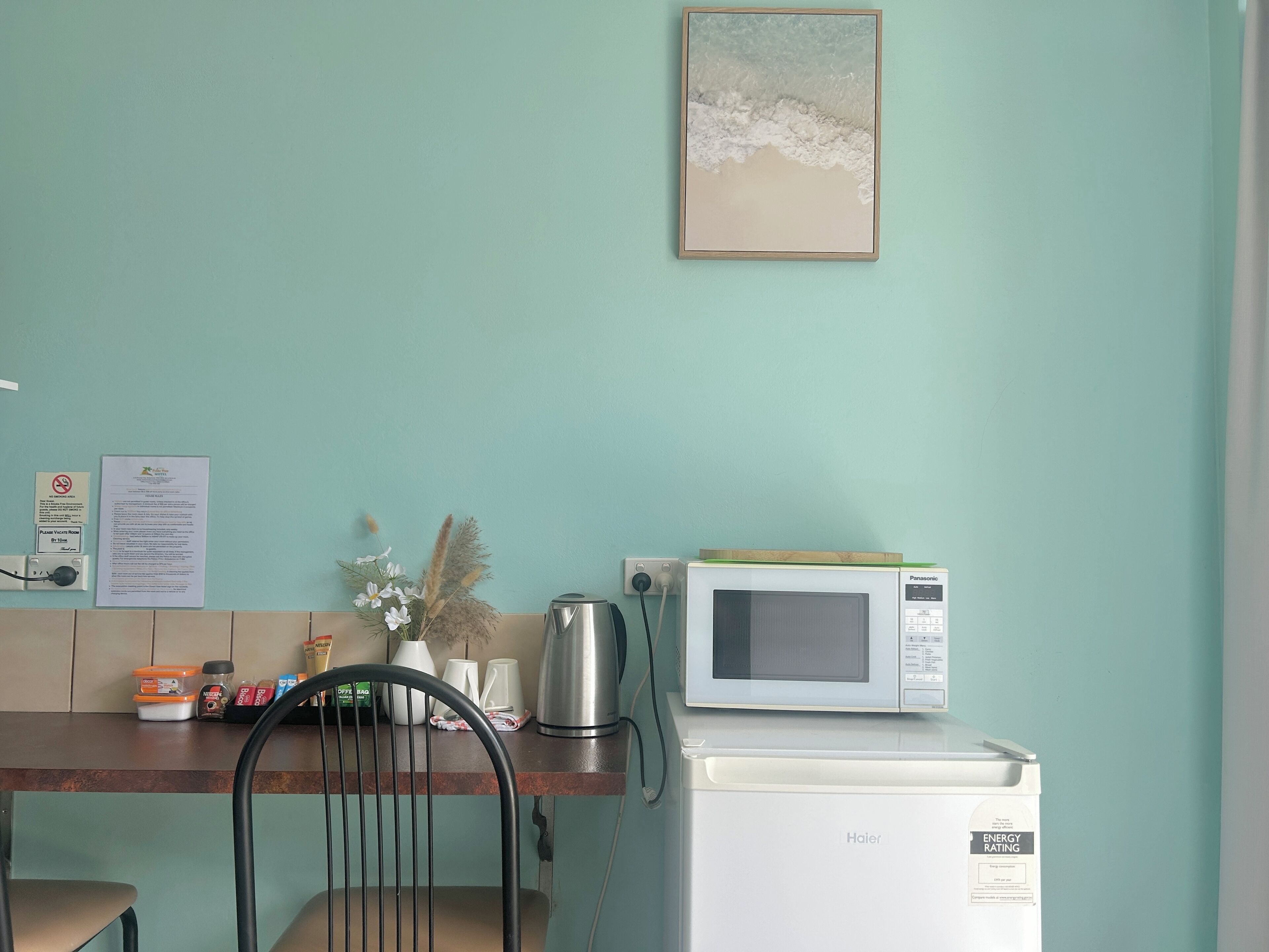 Una kitchenette con microondas, un refrigerador pequeño y una silla. Hay un cuadro colgado en la pared y un aviso.
