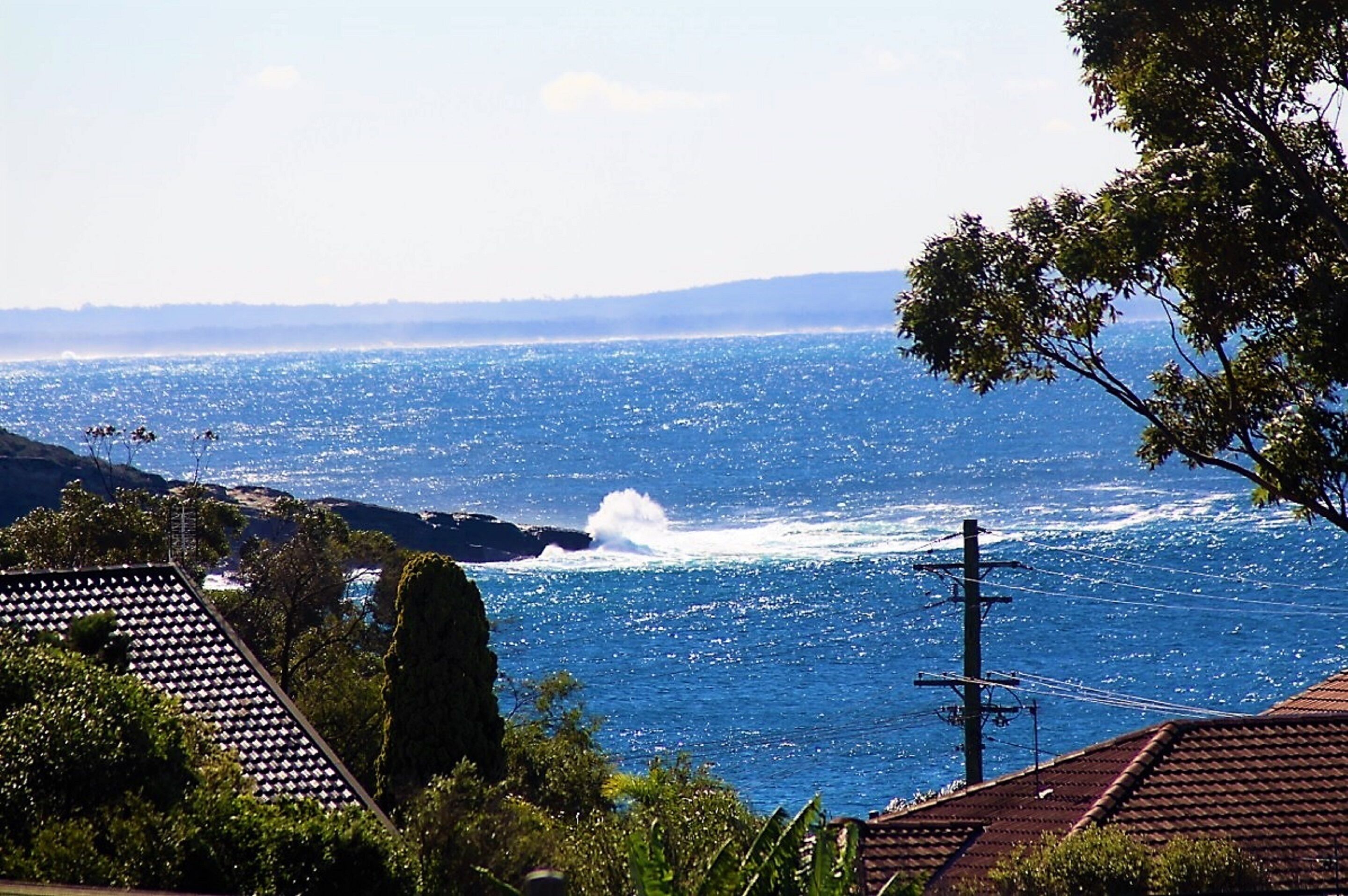 A coastal view with a clear sky, a rocky shoreline, and a building with a tiled roof.