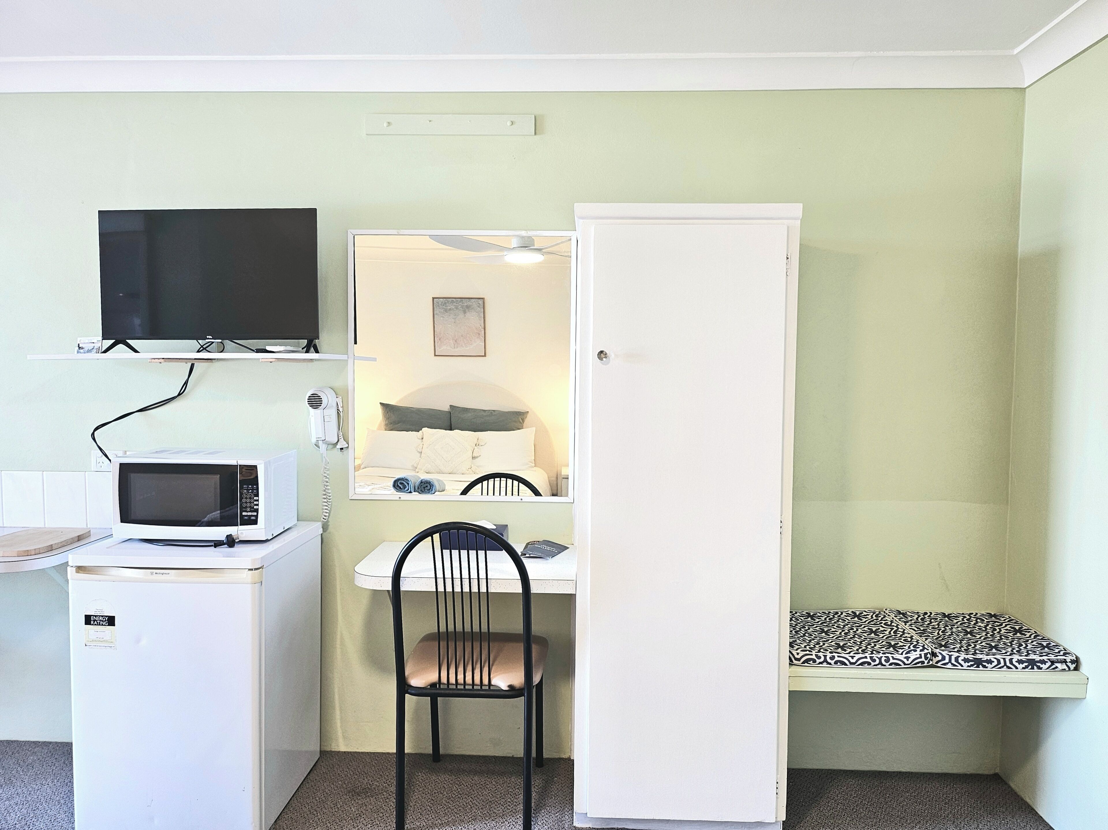 Quarto compacto com cama, escrivaninha e cadeira, micro-ondas, televisão e guarda-roupa.
