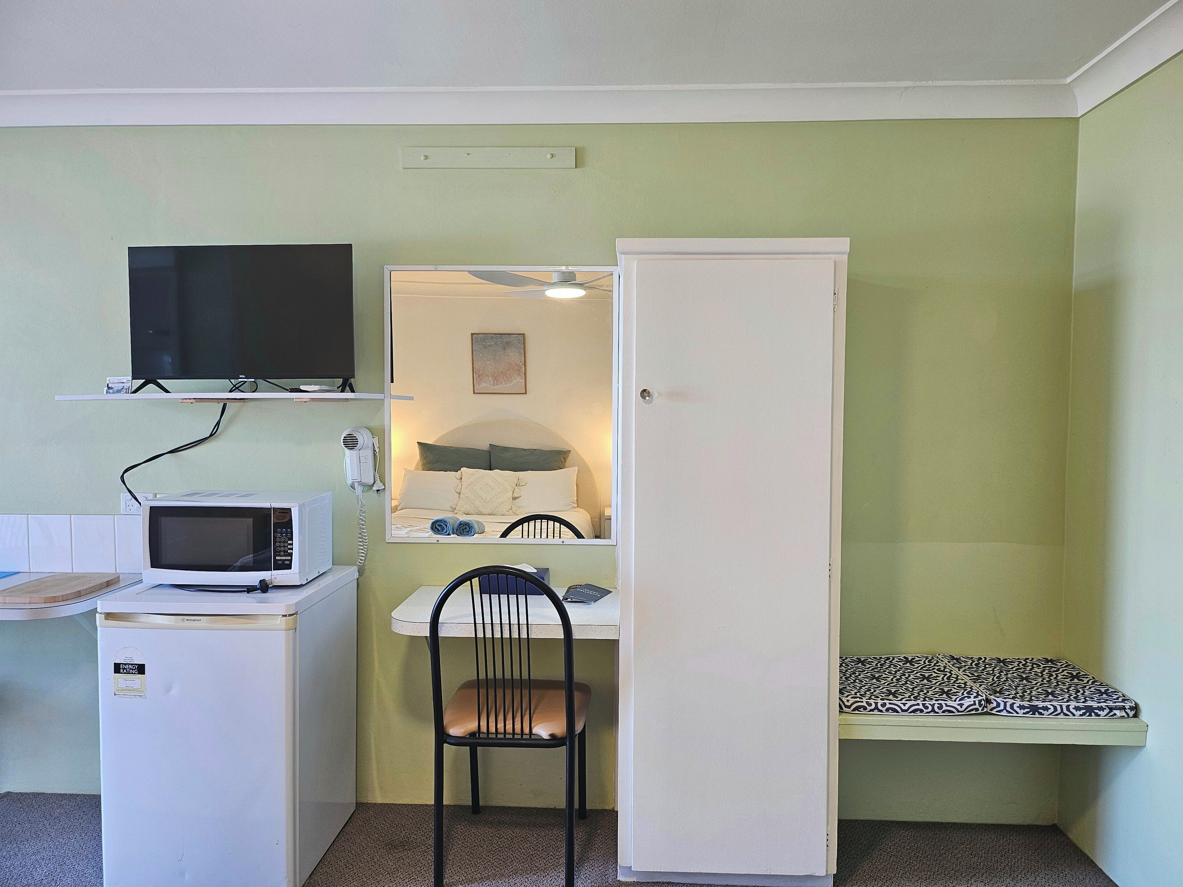 Une chambre de taille modeste comprenant un lit, un petit bureau avec une chaise, un four à micro-ondes, une télévision et une armoire.