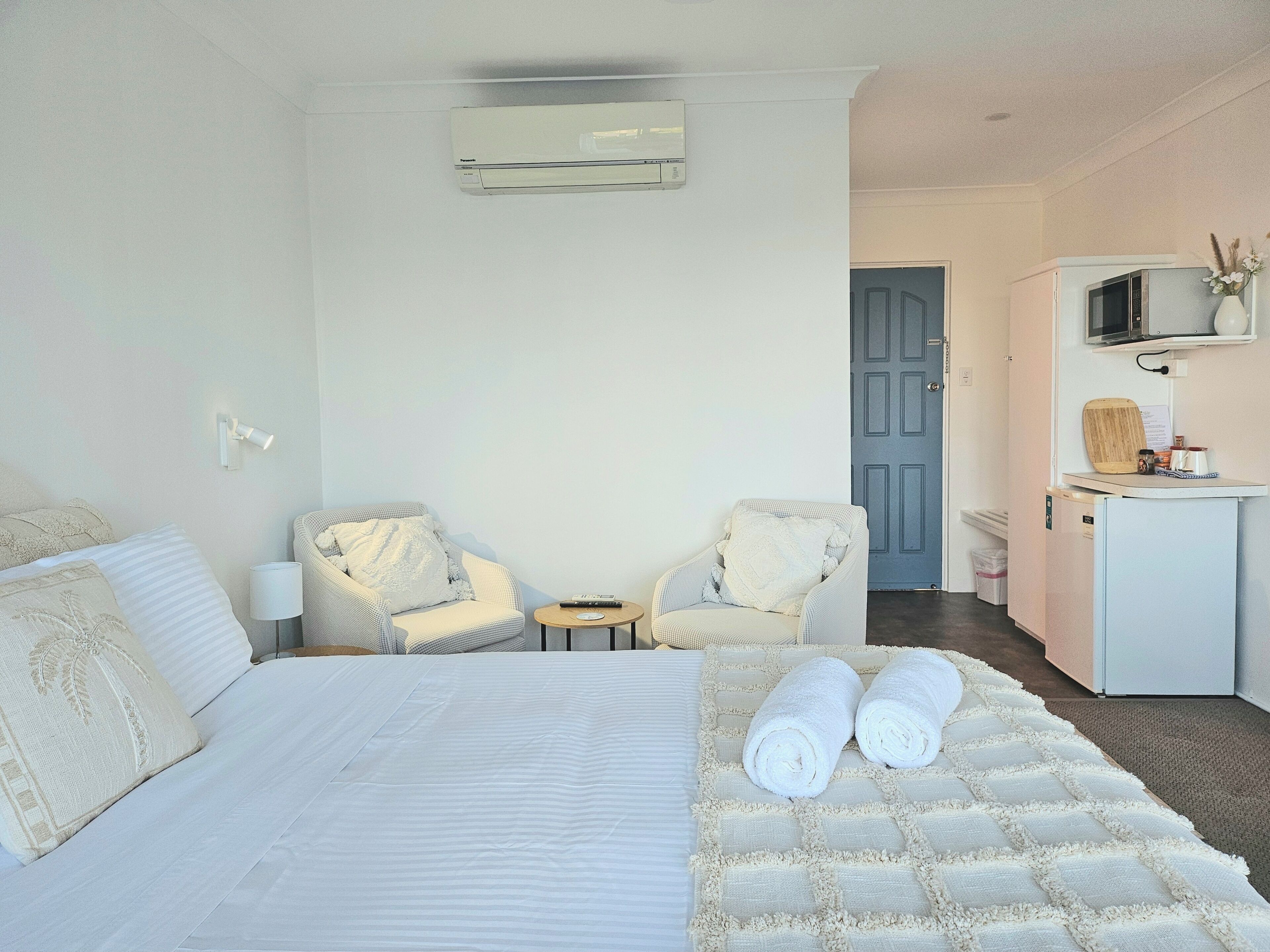 A hotel room with a bed, two armchairs, a small table, an air conditioning unit, a microwave, and a sink.