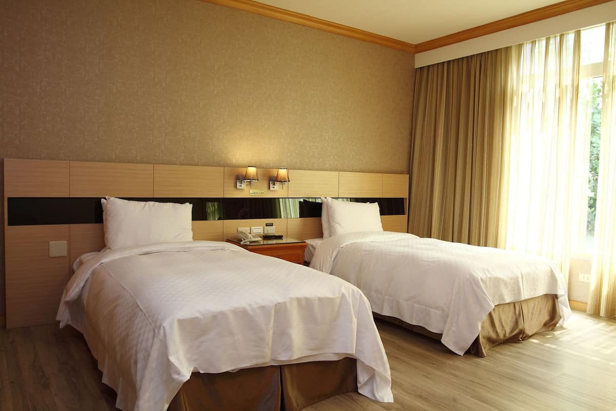 Quarto de hotel com duas camas, cabeceira de madeira, luminárias de cabeceira, mesa de cabeceira e janela com cortinas.