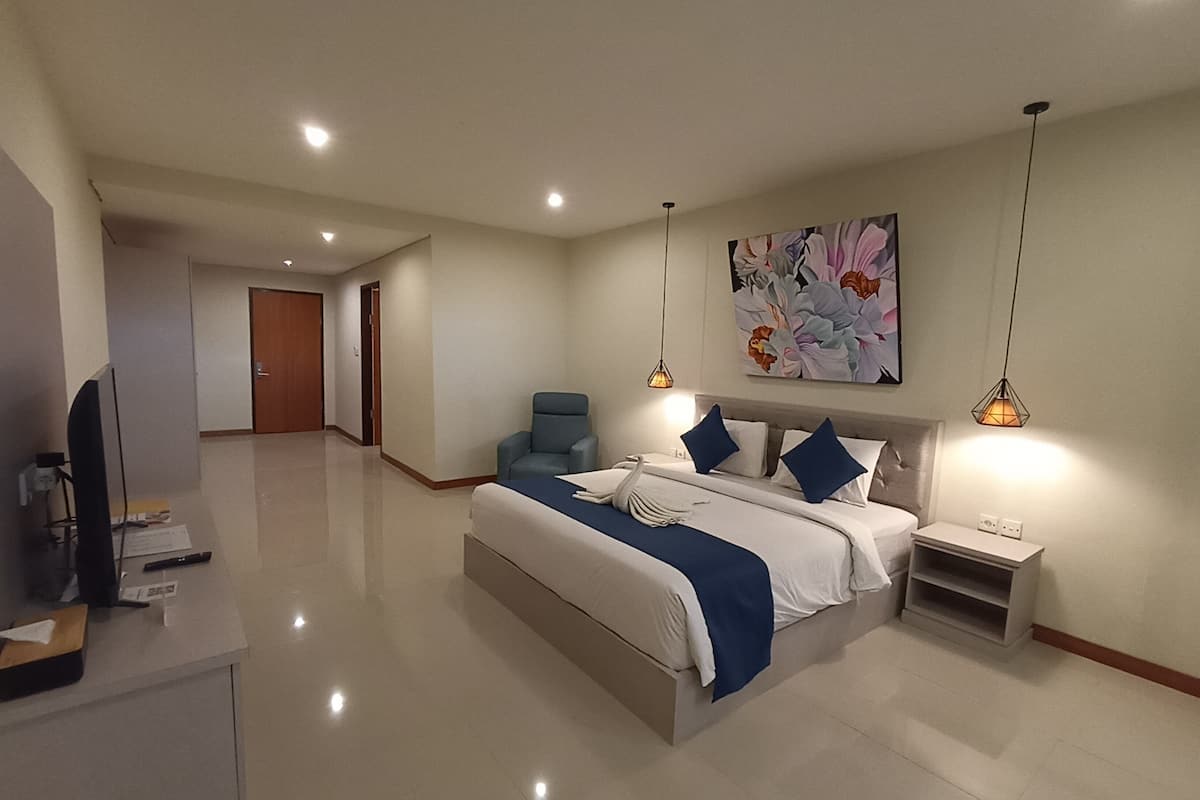 Ein modernes Hotelzimmer mit einem großen Bett, einem Schreibtisch, einem Sessel und einem Fernseher. An der Wand hängt ein Gemälde, und eine Deckenleuchte sorgt für Licht.