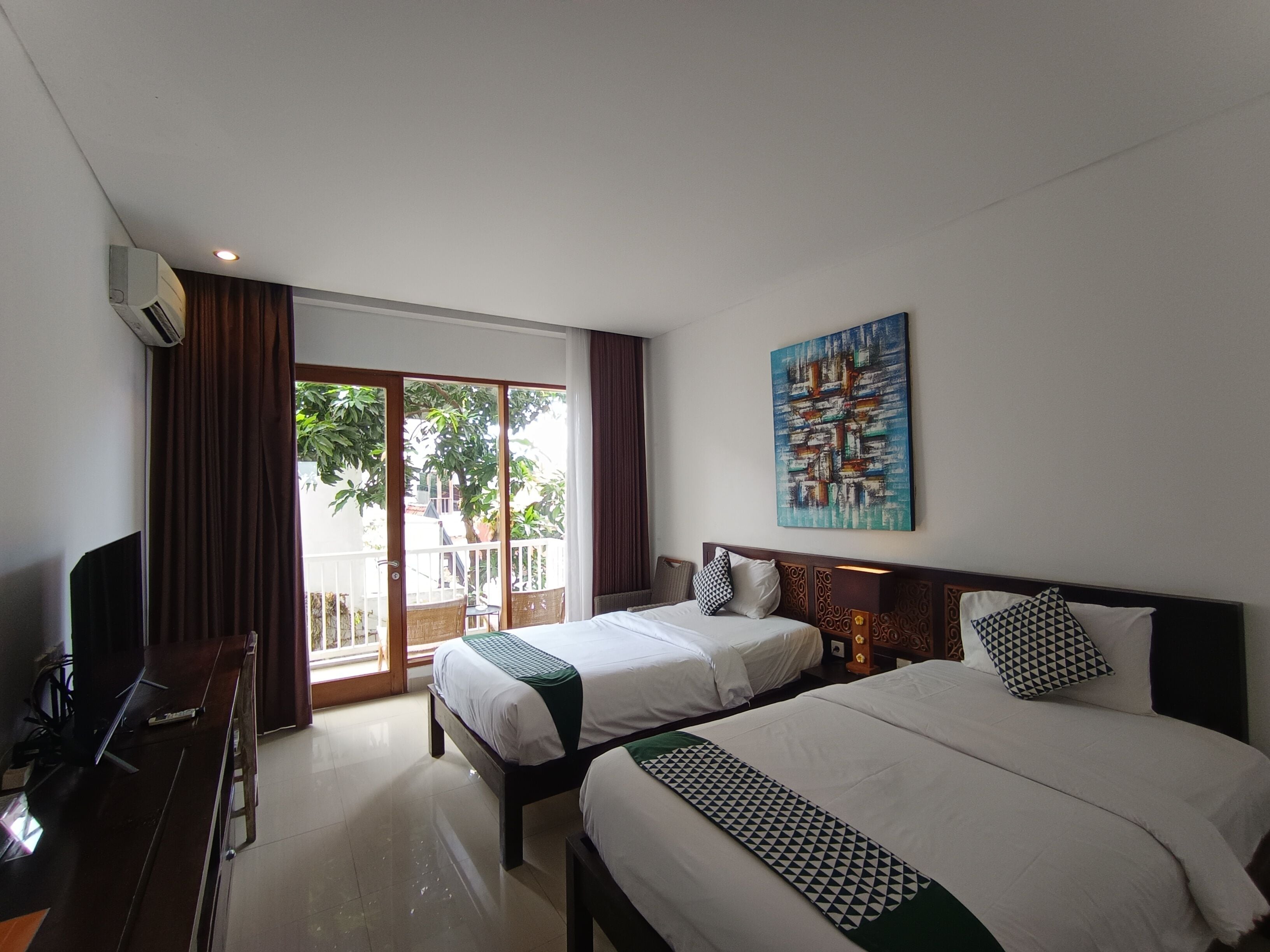 A hotel room with two beds, a desk, a TV, and a large window with a view of trees.